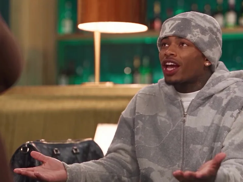 A man in a grey camouflage hoodie and beanie holds his hands out mid-argument beside a designer bag